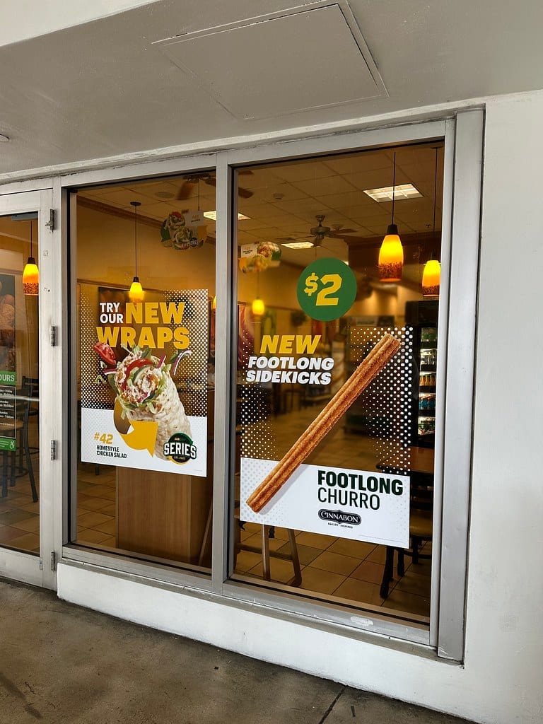 Subway storefront window with promotional menu signage and the dining area visible through the glass