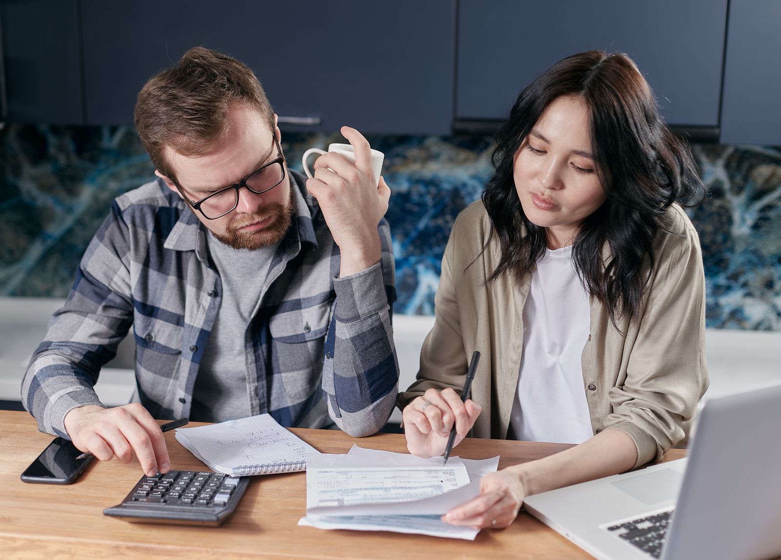 A couple at a kitchen table reviewing bills and a budget with a calculator and a laptop