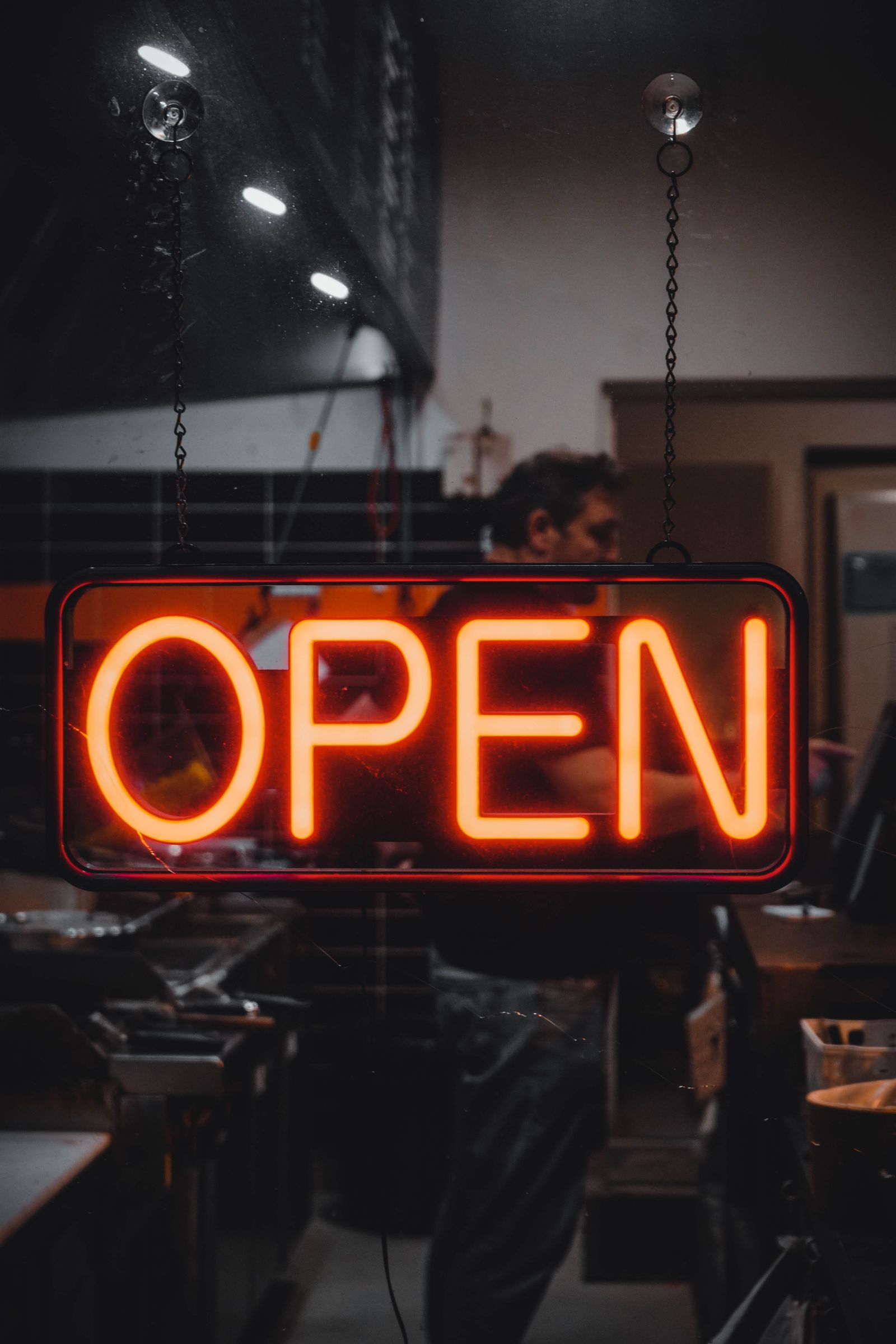 A glowing red OPEN neon sign in a shop window with the owner working behind it