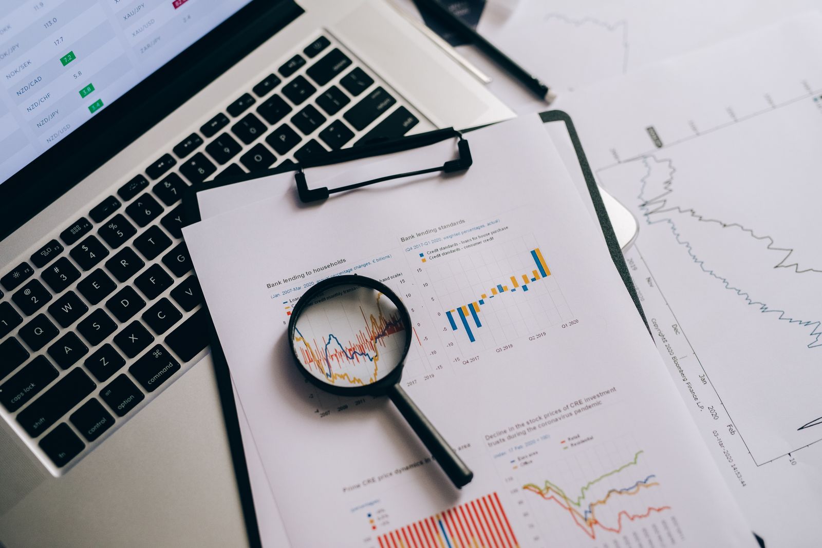 A magnifying glass over printed financial charts beside a laptop showing market data