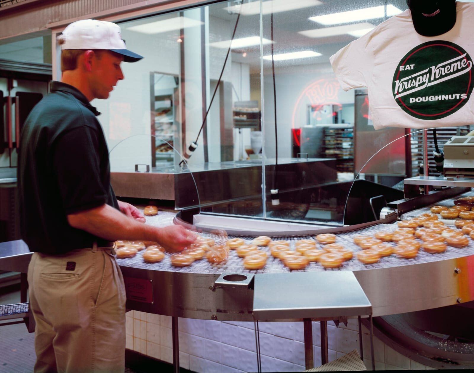 Krispy Kreme employee watching glazed doughnuts move down the production line conveyor inside a Hot Light Theater store