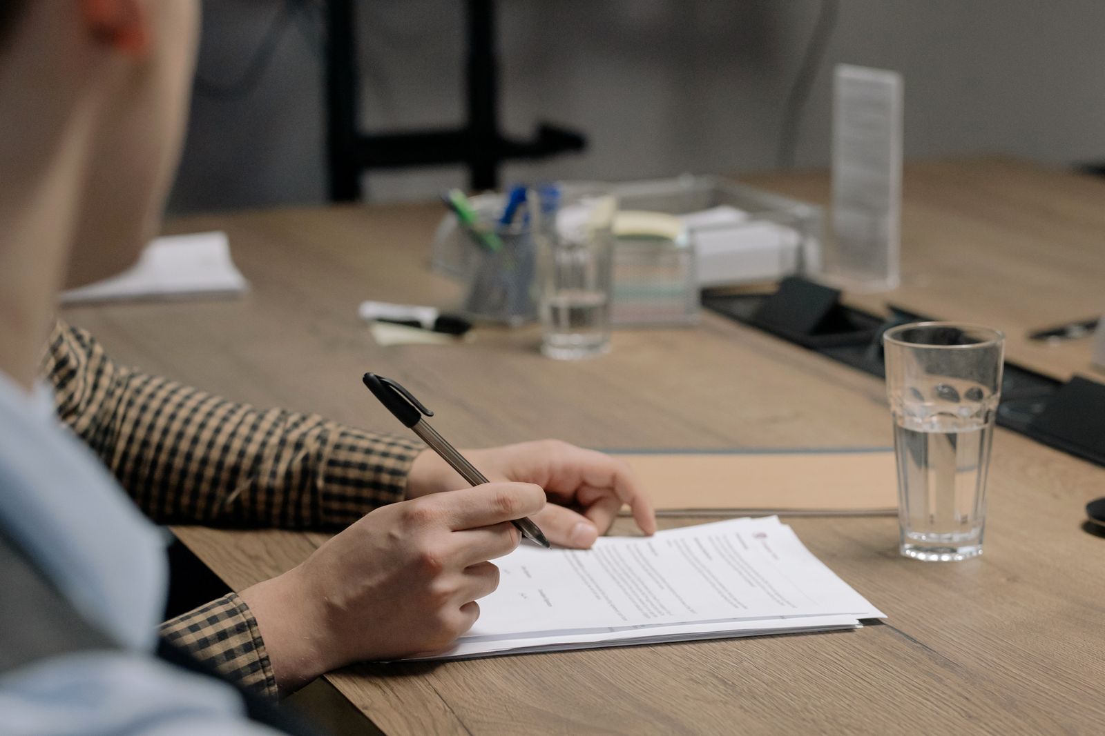 A person in a checked shirt signing a printed document with a ballpoint pen at a wooden office table